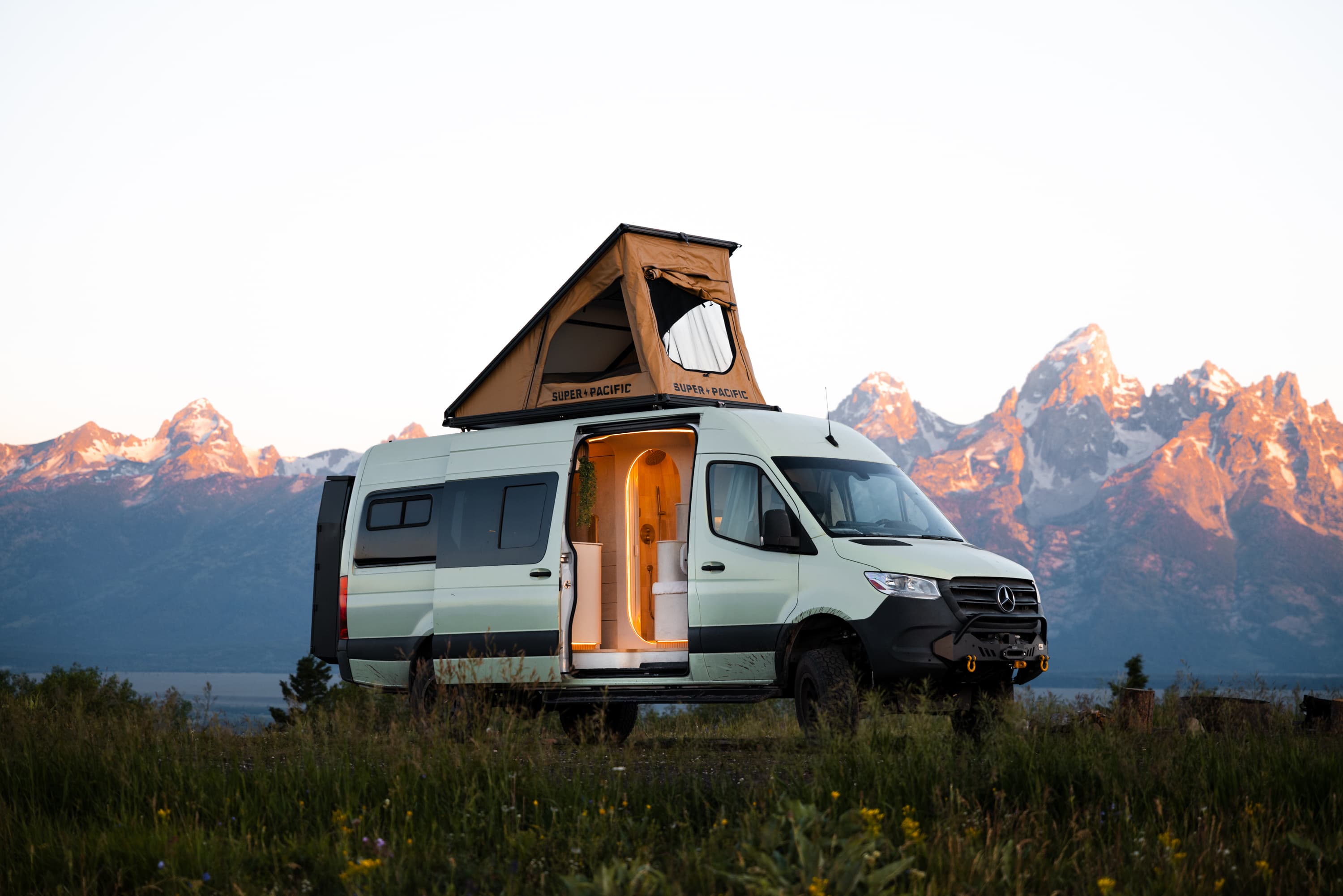The Flow, a custom Mercedes Sprinter, parked below the Tetons at sunset with its pop-top raised and interior lit.