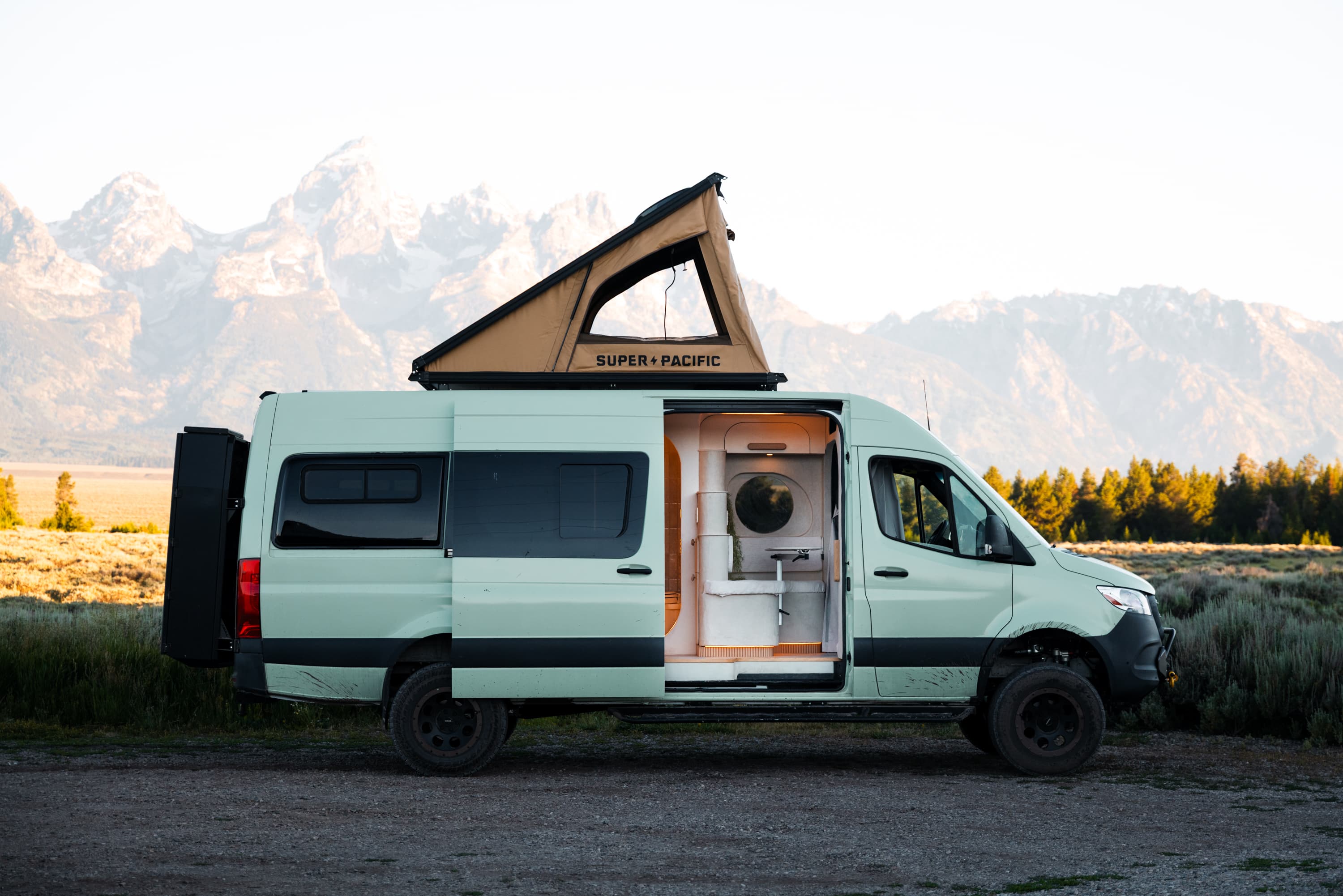 The Flow in profile with its sliding door open and pop-top raised, the sculpted interior visible against the Tetons.